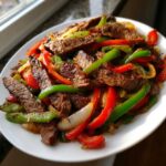 Close-up of sizzling steak fajitas featuring strips of seasoned steak, red and green bell peppers, and onions on a white plate.