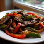 Close-up of sizzling steak fajitas with colorful strips of red and green bell peppers and onions on a white plate.