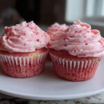 Two perfectly frosted strawberry cupcakes with pink frosting flecked with red pieces, sitting on a white plate.