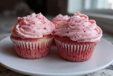 Two perfectly frosted strawberry cupcakes with pink frosting flecked with red pieces, sitting on a white plate.
