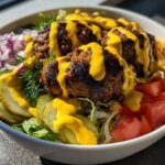Close-up of a white bowl filled with a cheeseburger bowl featuring seasoned meat patties, pickles, tomatoes, onions, and bright yellow sauce.