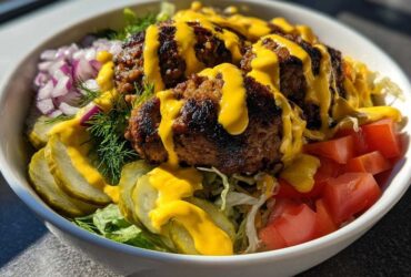 Close-up of a white bowl filled with a cheeseburger bowl featuring seasoned meat patties, pickles, tomatoes, onions, and bright yellow sauce.
