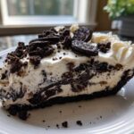 A generous slice of creamy, no-bake Oreo pie with a dark crust, topped with whipped cream and cookie pieces.