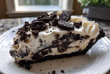 A generous slice of creamy, no-bake Oreo pie with a dark crust, topped with whipped cream and cookie pieces.
