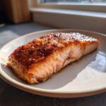 A perfectly cooked, flaky fillet of air fryer salmon with a caramelized, seasoned crust resting on a light-colored plate.