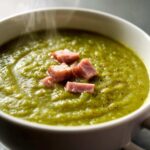 Close-up of a steaming bowl of thick, vibrant green pea soup topped with diced ham pieces and black pepper.