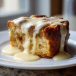 A close-up of a square slice of golden brown bread pudding topped with a thick vanilla cream sauce.