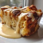 A close-up of a rich, square slice of bread pudding topped with a drizzle of vanilla sauce and sitting in a pool of custard.