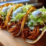 Three hard-shell chicken tacos filled with shredded seasoned chicken, lettuce, and sour cream, sitting on a wooden board.