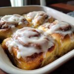 Four freshly baked cinnamon roll pastries covered in thick, white cream cheese frosting in a baking dish.