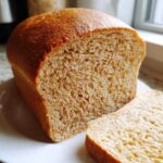 A freshly baked loaf of whole wheat bread, partially sliced, resting on a white plate.
