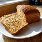 A golden brown loaf of whole wheat bread, partially sliced, resting on a white plate in natural light.
