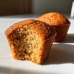 Close-up of a banana protein muffin with a bite taken out, showing the fluffy interior next to a whole muffin.