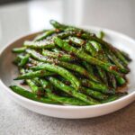 A close-up of bright green, blistered chinese green beans with garlic sauce piled high in a white bowl.