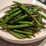 A close-up of blistered chinese green beans with garlic sauce piled high on a light ceramic plate.