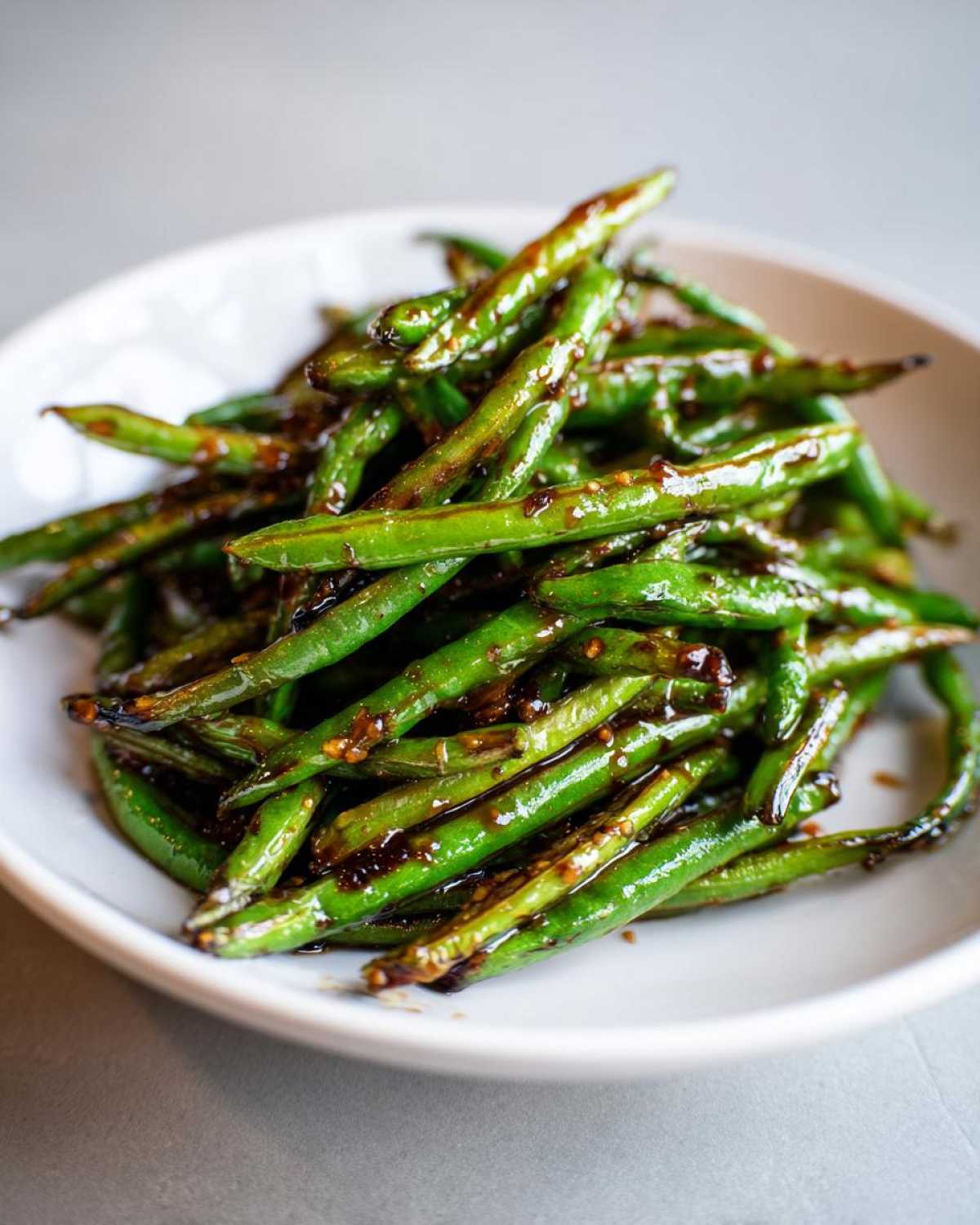 A close-up of blistered chinese green beans with garlic sauce piled high in a white bowl.