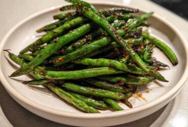 A close-up of blistered chinese green beans with garlic sauce piled high on a light ceramic plate.
