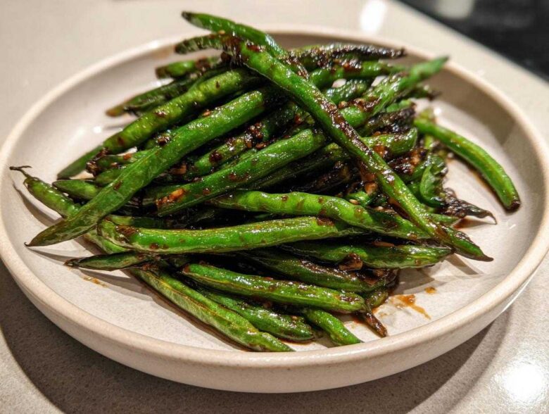 A close-up of blistered chinese green beans with garlic sauce piled high on a light ceramic plate.