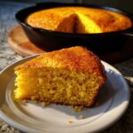 A golden slice of buttermilk cornbread served on a white plate with the rest of the loaf visible in a cast iron skillet.
