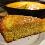 A close-up of a golden slice of moist buttermilk cornbread served on a white plate, with the rest of the cornbread in the cast iron skillet in the background.