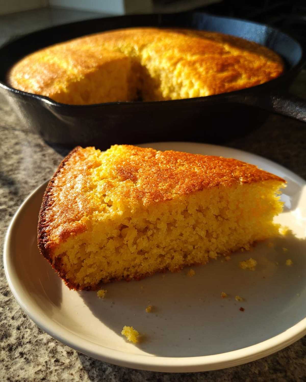 A golden slice of buttermilk cornbread served on a plate with the rest of the cornbread visible in the cast iron skillet behind it.