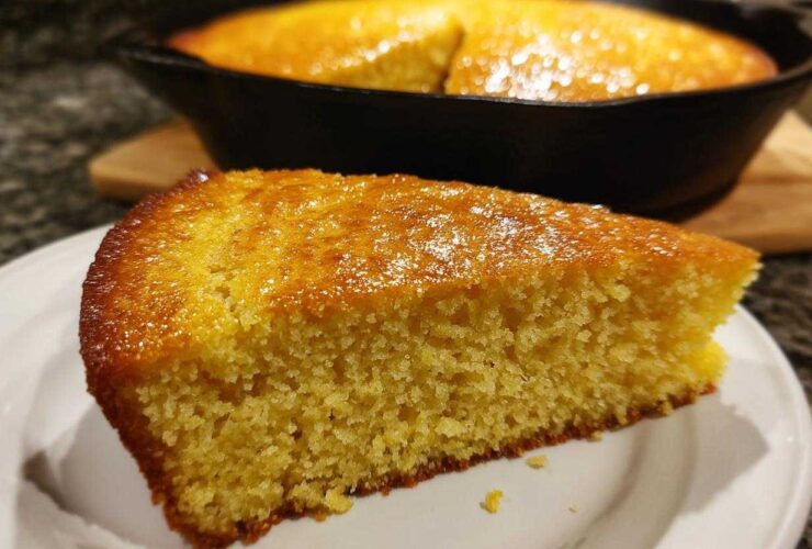 A close-up of a golden slice of moist buttermilk cornbread served on a white plate, with the rest of the cornbread in the cast iron skillet in the background.