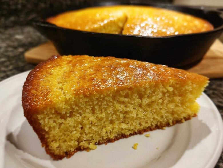 A close-up of a golden slice of moist buttermilk cornbread served on a white plate, with the rest of the cornbread in the cast iron skillet in the background.