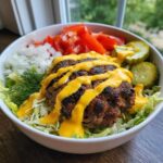 A vibrant cheeseburger bowl featuring a seasoned patty topped with yellow cheese sauce, served over lettuce with diced onions, tomatoes, and pickles.