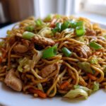 A close-up of a serving of homemade chicken chow mein recipe, featuring noodles, chicken pieces, cabbage, and green onions.