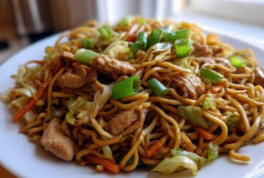 A close-up of a serving of homemade chicken chow mein recipe, featuring noodles, chicken pieces, cabbage, and green onions.
