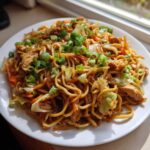 Close-up of a white plate piled high with savory chicken chow mein recipe, garnished with fresh green onions.