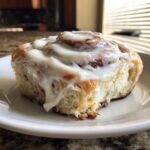 A single cinnamon roll generously topped with thick, white cream cheese frosting on a white plate.