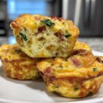 A stack of three golden-brown cottage cheese egg cups starbucks style, one cut open showing the fluffy interior with bacon and spinach.