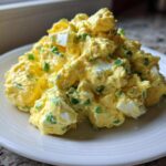 A generous mound of creamy, yellow egg salad recipe mixed with chopped green onions, served on a white plate.