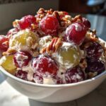 Close-up of a white bowl filled with creamy grape salad, featuring red, green grapes, and chopped pecans.