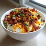 A white bowl filled with creamy Million Dollar Dip, topped generously with shredded cheddar cheese, crispy bacon bits, and sliced green onions.