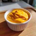 A small white ramekin filled with thick, bright orange nacho cheese sauce, sitting on a wooden surface.