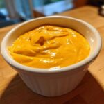 A close-up of thick, vibrant orange nacho cheese sauce served in a small white fluted ramekin on a wooden surface.