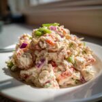 A mound of creamy seafood salad mixed with red onion and celery, garnished with green onion on a white plate.
