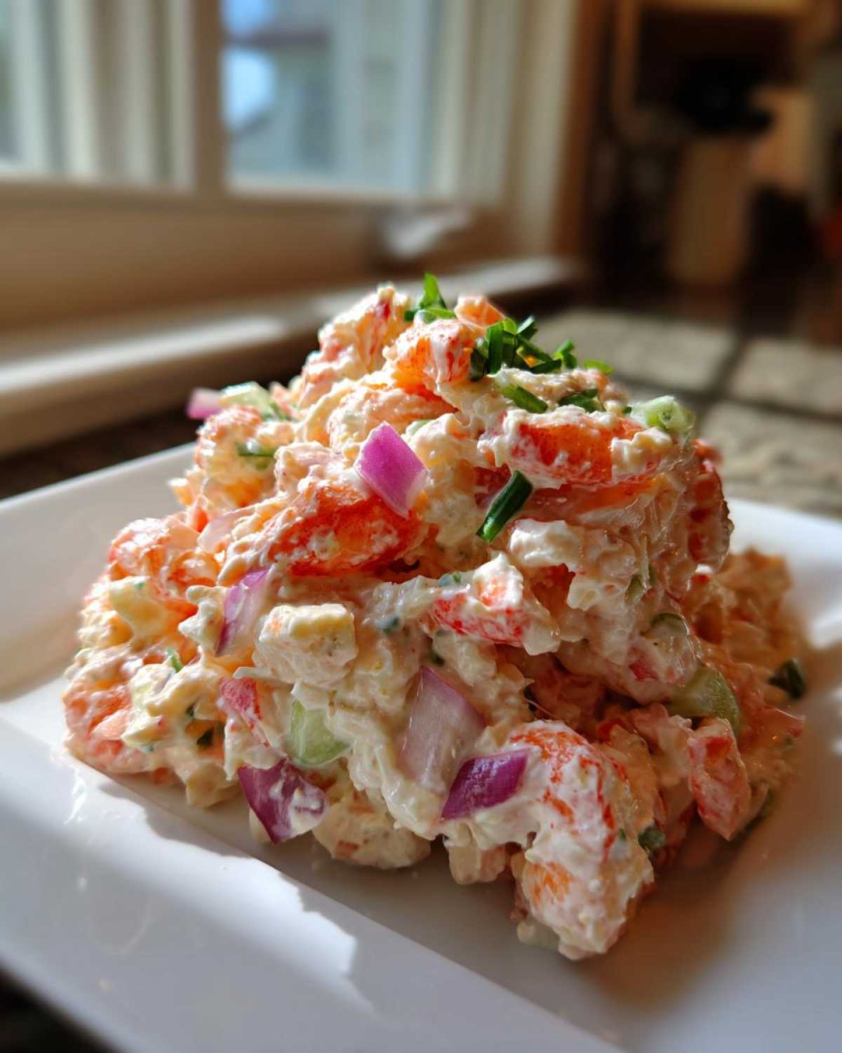 A close-up of a creamy seafood salad featuring shrimp, red onion, and chives, served on a white square plate.