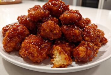 A mound of crispy, glazed orange chicken recipe pieces piled high on a white plate, showing a bite taken out.