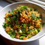 A close-up of a bowl filled with crispy rice salad mixed with bright green edamame beans and fresh mint leaves.