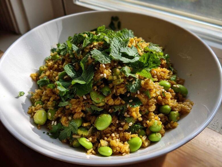 A white bowl filled with golden brown crispy rice salad mixed with bright green edamame and topped generously with fresh mint and cilantro.
