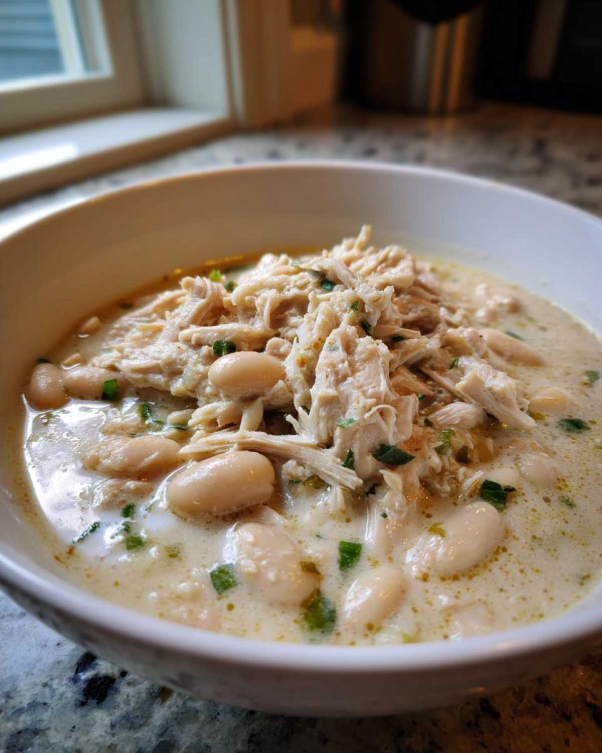 A close-up of a white bowl filled with creamy crockpot white chicken chili, topped with shredded chicken and green onions.
