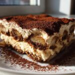 A perfect slice of classic tiramisu recipe showing layers of cream and coffee-soaked ladyfingers, heavily dusted with cocoa powder.