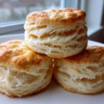 Three golden-brown, flaky buttermilk biscuits stacked on a white plate near a window.