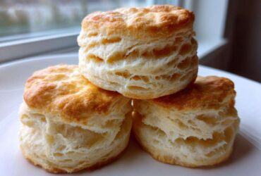 Three golden-brown, flaky buttermilk biscuits stacked on a white plate near a window.