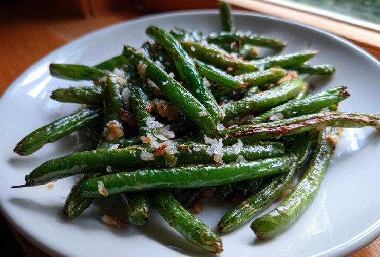 Close-up of bright green beans seasoned with coarse salt and browned bits, served on a white plate.