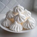 A close-up of several perfectly piped, bright white meringue cookies stacked on a white plate.