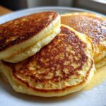 Close-up of three fluffy almond flour pancakes stacked on a white plate, drizzled with syrup.